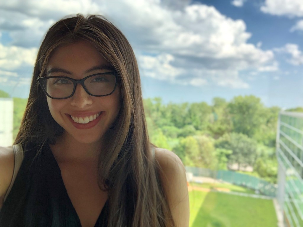 A woman with brown hair and glasses smiles at the camera in front of a background of trees behind a field.