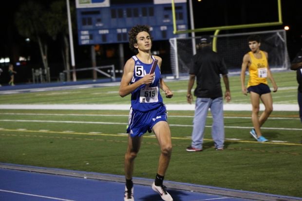 A boy in Flying L track uniform carries a relay baton while running on the track.