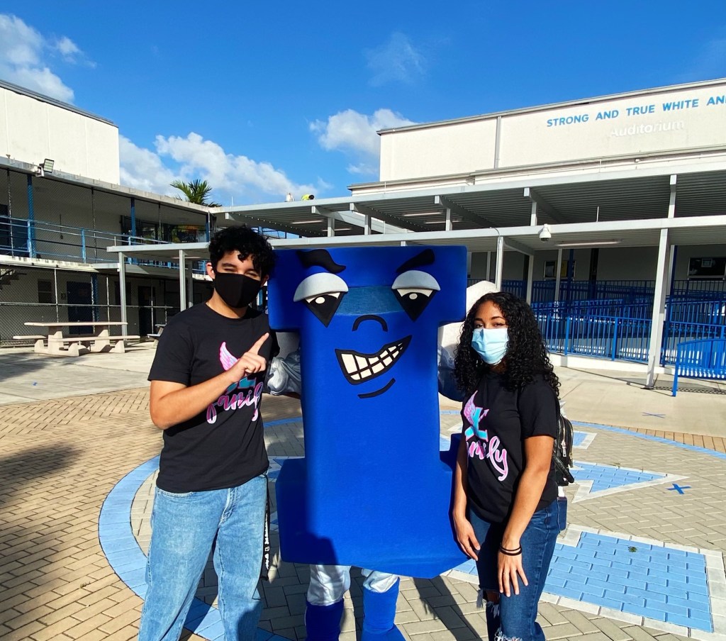 A boy and a girl stand on each side of a person in a winged blue L costume in front of the school auditorium.