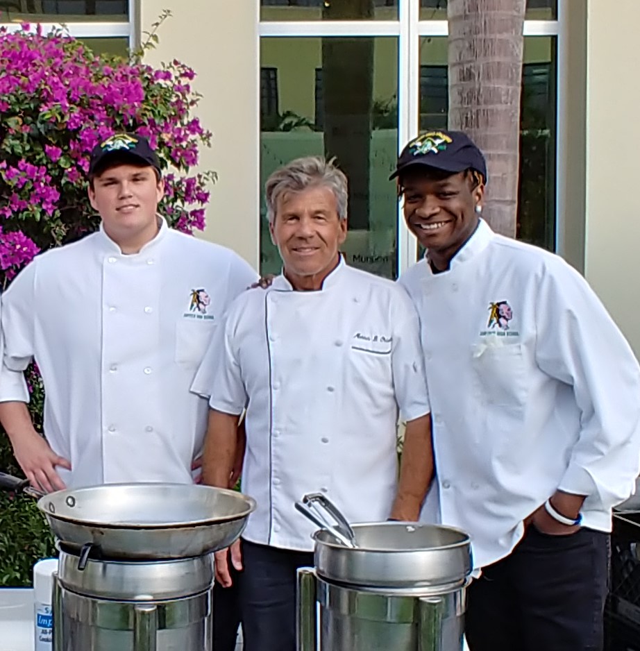 Three men in chef's jackets stand outside behind a table with cooking implements.