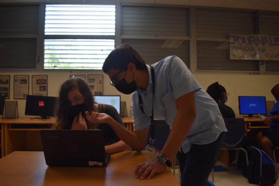 A boy leaning over the side of a desk counsels another sitting with a laptop.
